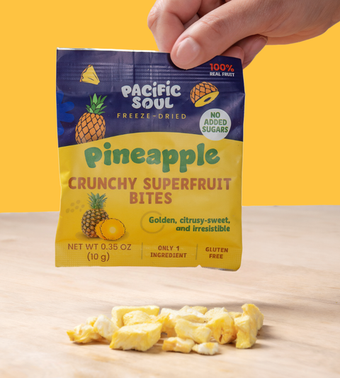 Hand holding a package of Pacific Soul Pineapple Crunchy Superfruit Bites on a wooden surface with a yellow background.