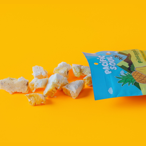 A bag of Pacific Soul Pineapple supefruit bites sits on a bright yellow background with several pieces of freeze-dried pineapple spilled out in front. The bag is blue with a pineapple illustration and the brand name 'Pacific Soul' is clearly visible.